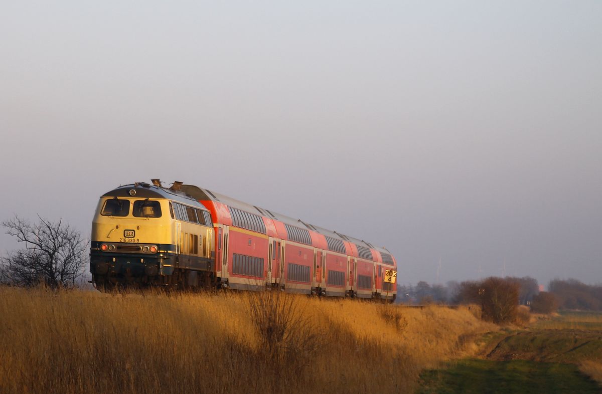 Die seit dem 25.02.2022 im AW Cottbus wieder blau-beige lackierte DB 218 330  Konrad  des Bw Kiel schiebt ihren Doppelstockzug im letzen Licht nach Husum, hier am Bü Triangel 06.03.2025